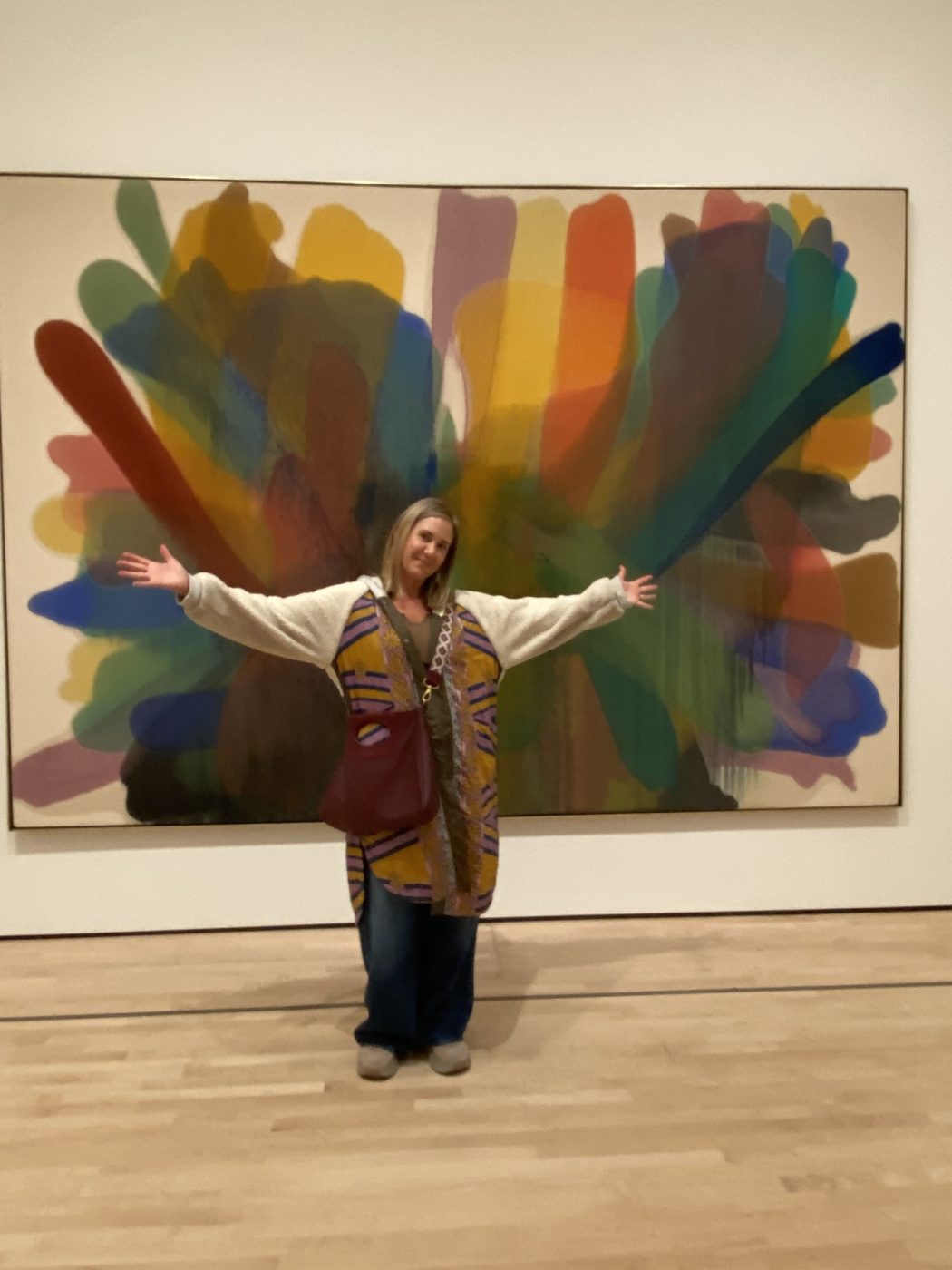Amy with arms outstretched in front of a large rainbow painting at a museum.