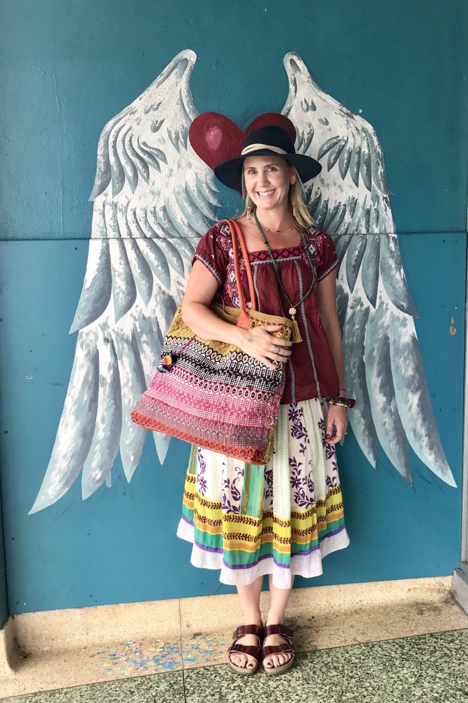 Amy standing in front of a painted pair of angel wings, carrying a woven bag.