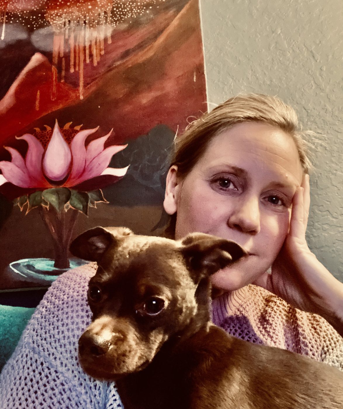 Amy resting her chin on her hand with her dog, in front of a lotus painting.