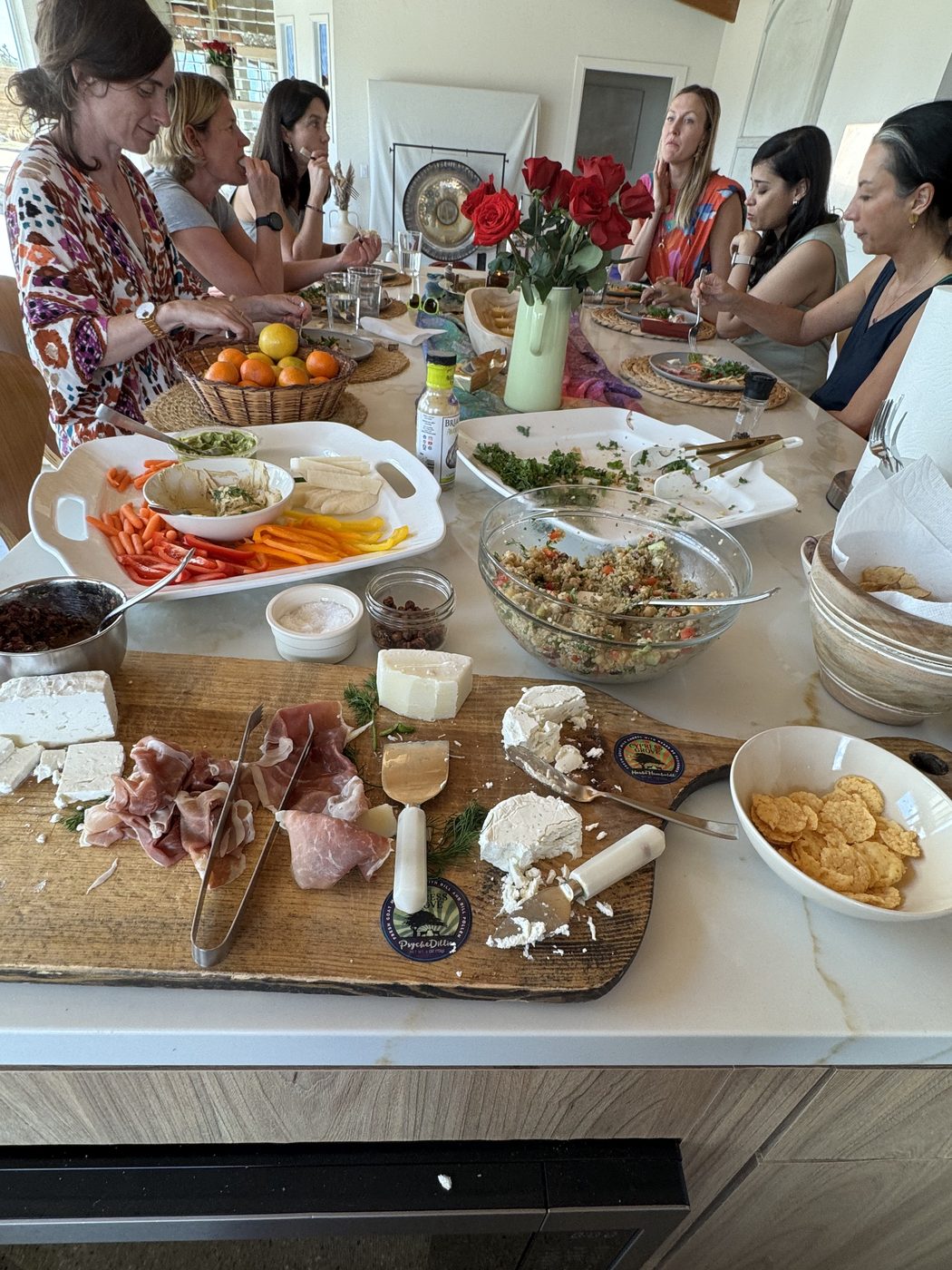 Women gathered around a kitchen island laid with a grazing board, salads and flowers.
