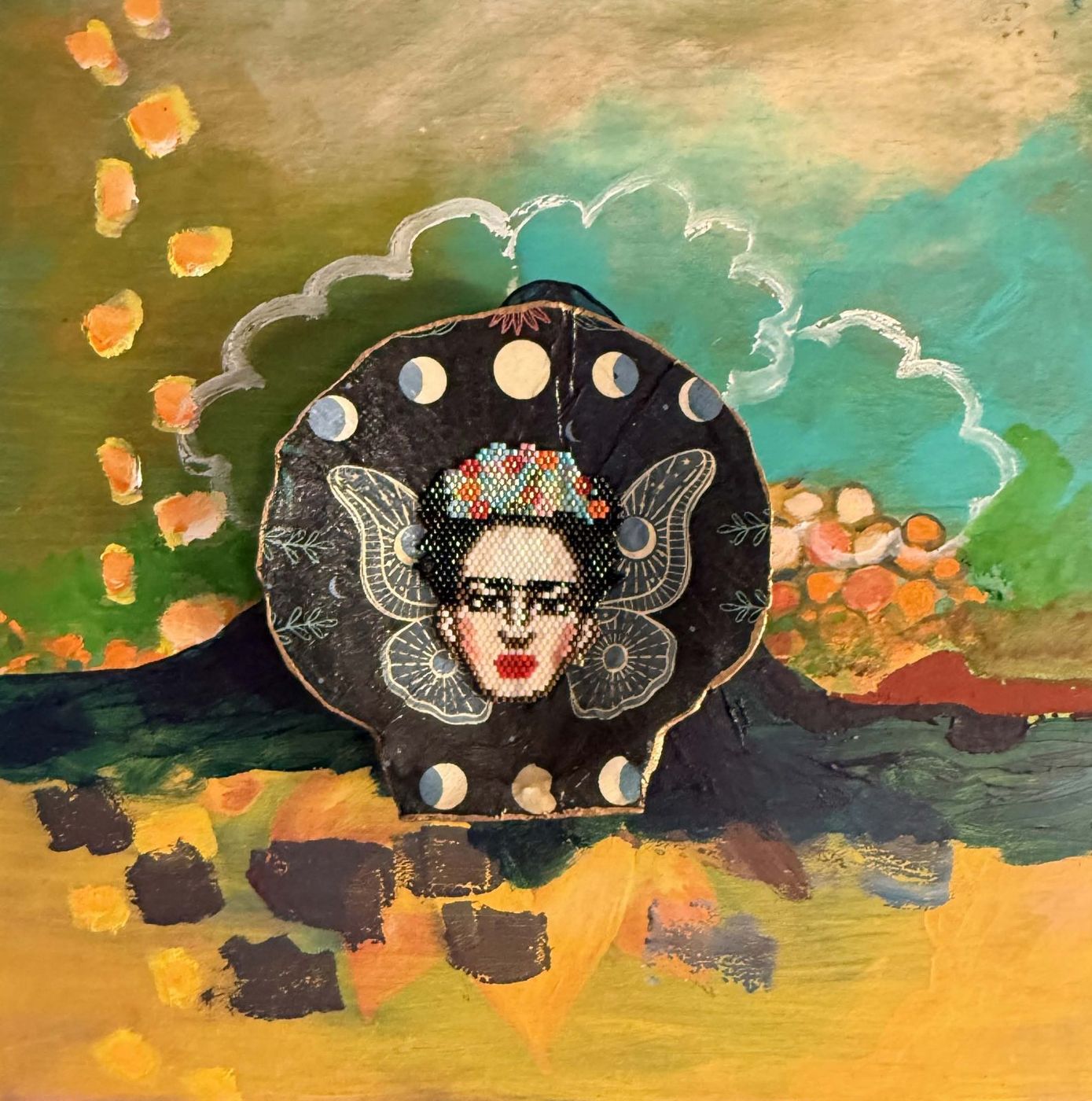Painting with a beaded Frida Kahlo face set in a dark moon-phase disc, over a green and gold landscape.