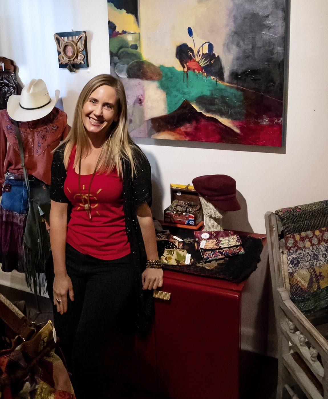 Amy smiling in the Goddess Gatherings shop beside a painting and vintage hats.