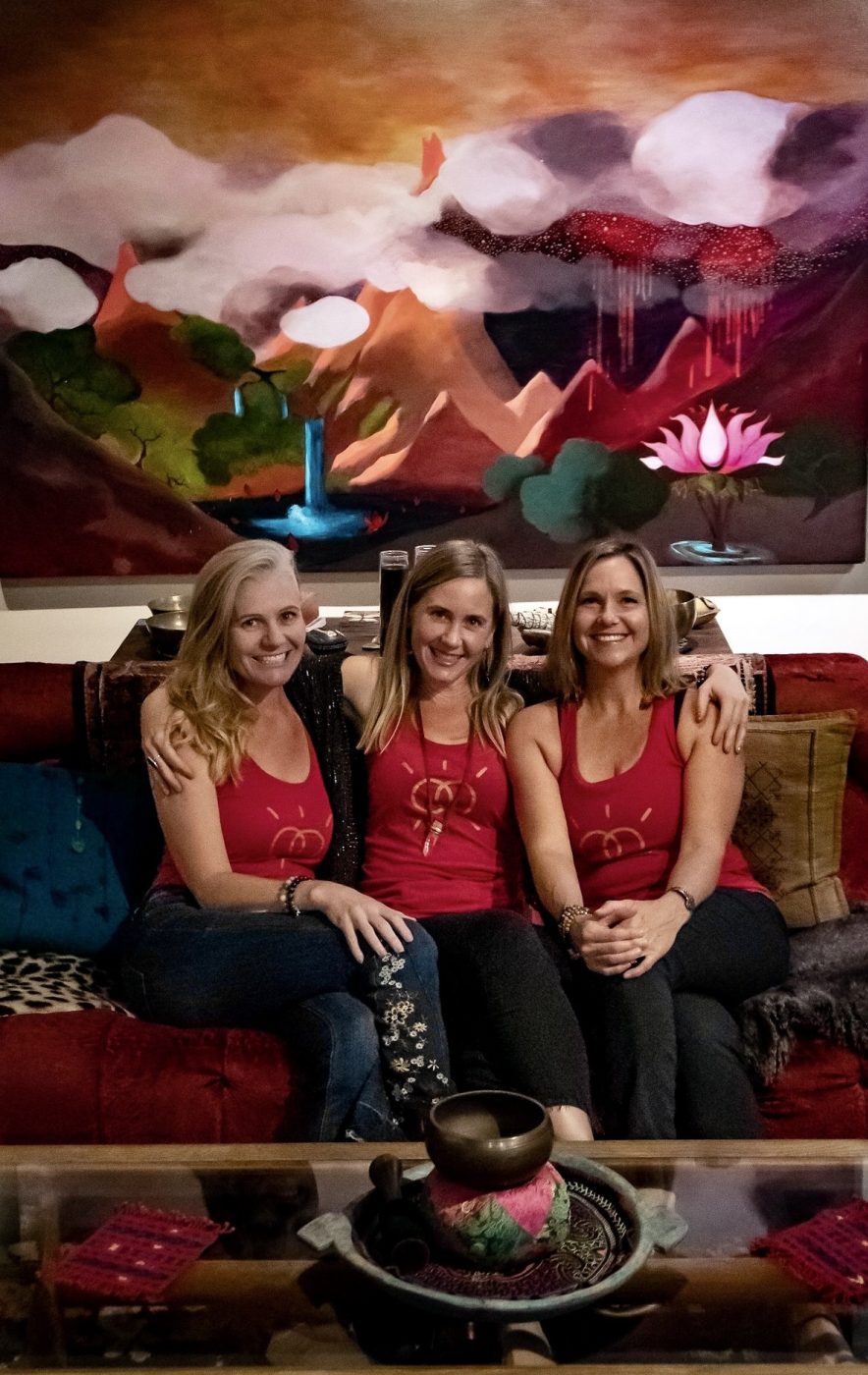 Three women in matching red tops on a sofa beneath the red mountain painting.