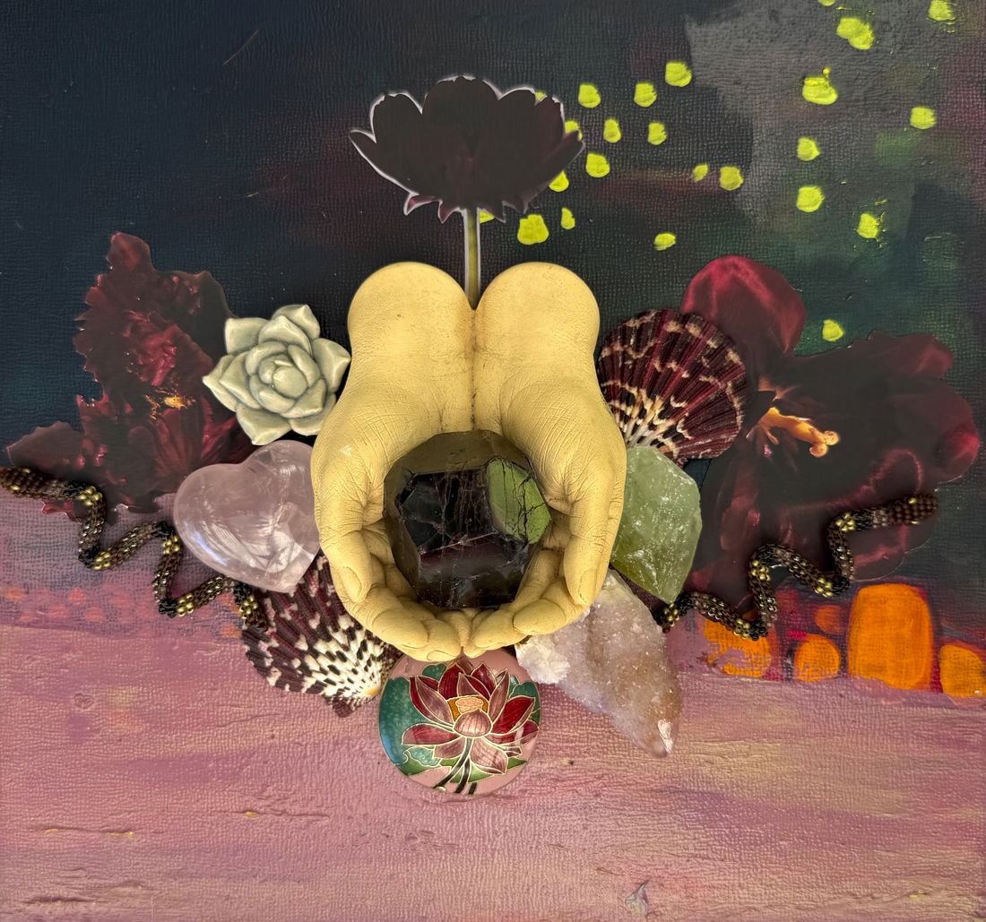 Sculpted cupped hands holding a dark crystal on a painted board, surrounded by shells, flowers, a rose quartz heart and a lotus medallion.
