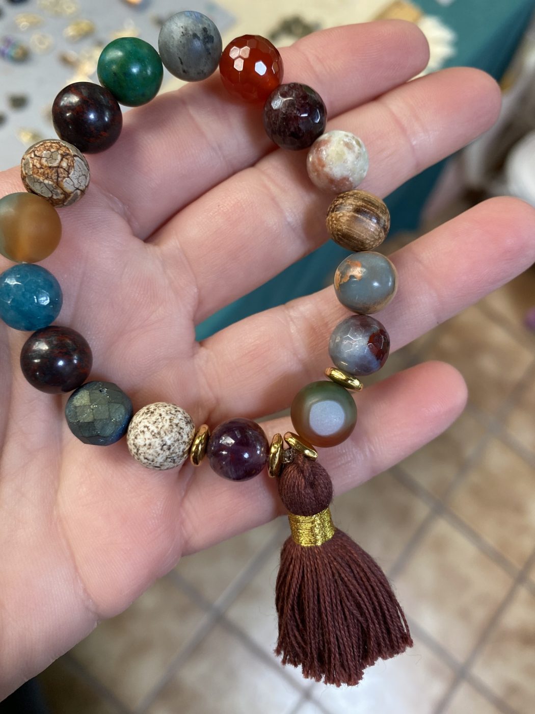 A hand holding a mixed agate bracelet with a brown tassel.