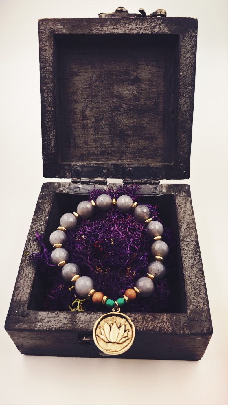 A grey bead bracelet with a brass lotus charm in a small wooden box.