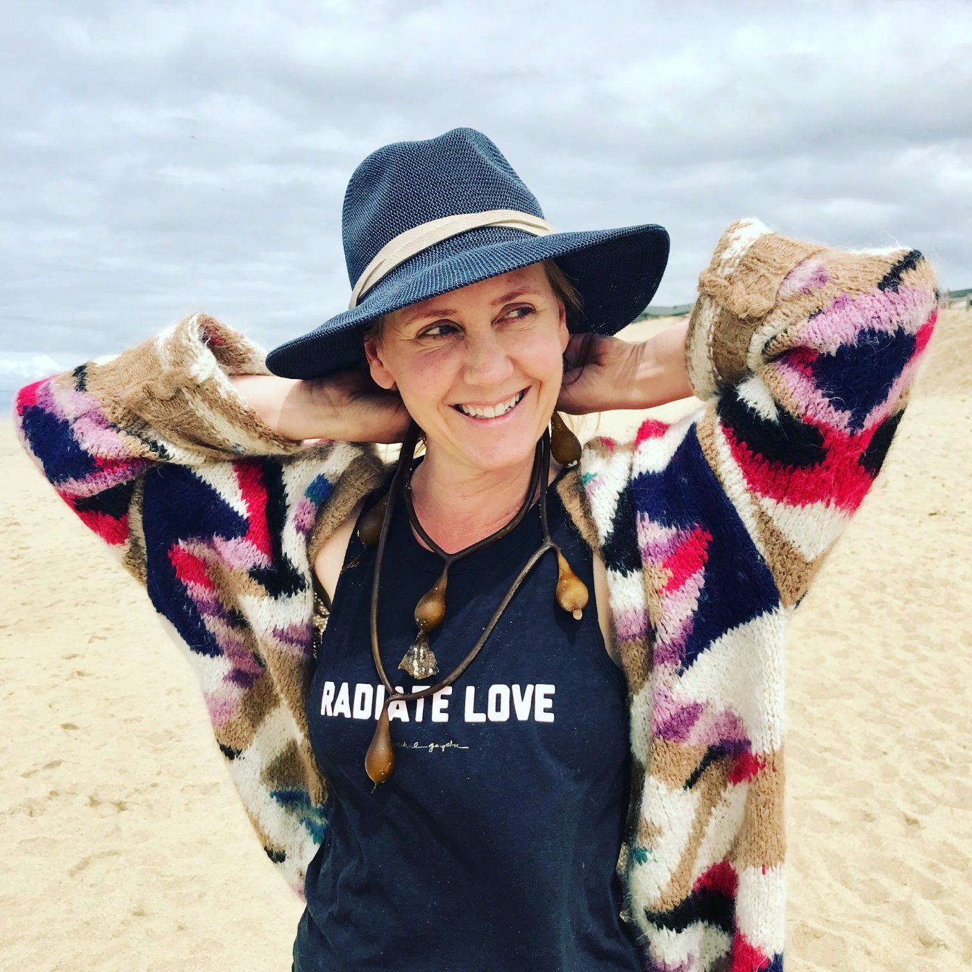 Amy on the beach in a hat and patterned cardigan, wearing a Radiate Love shirt.