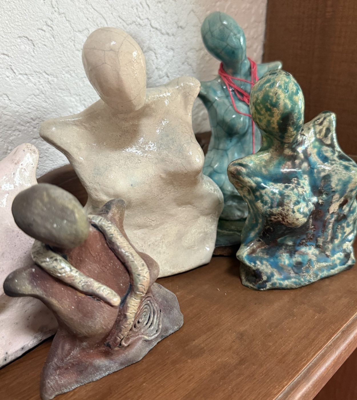 Four ceramic goddesses on a shelf: a cream crackle-glazed torso, a turquoise figure wrapped in red thread, a speckled blue-green figure and a brown figure with a coiled snake.