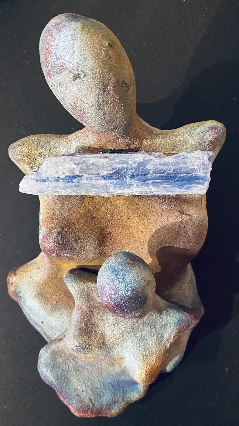 A raku goddess with a blue kyanite blade across her shoulders and a small sphere at her lap, in iridescent tan, blue and violet.