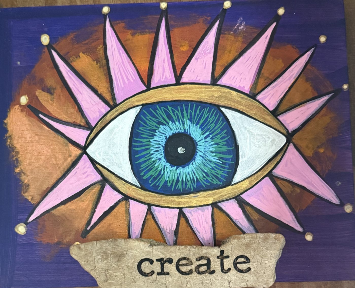 A painted eye with pink star rays on a purple and copper board, above a piece of driftwood stamped create.