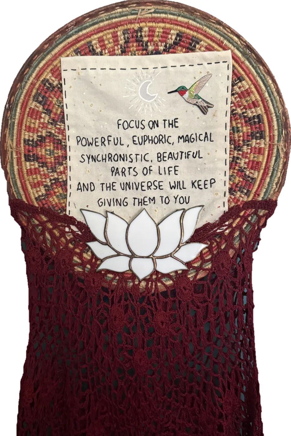 A woven basket with a hand-lettered cloth reading Focus on the powerful, euphoric, magical, synchronistic, beautiful parts of life and the universe will keep giving them to you, with a moon, a hummingbird, a stained glass lotus and deep red crochet.