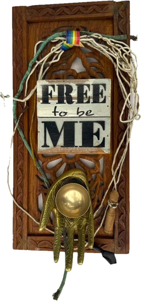 A carved wood panel with old wire, a rainbow ribbon, a brass hand holding a bowl, and a sign reading Free to be Me.