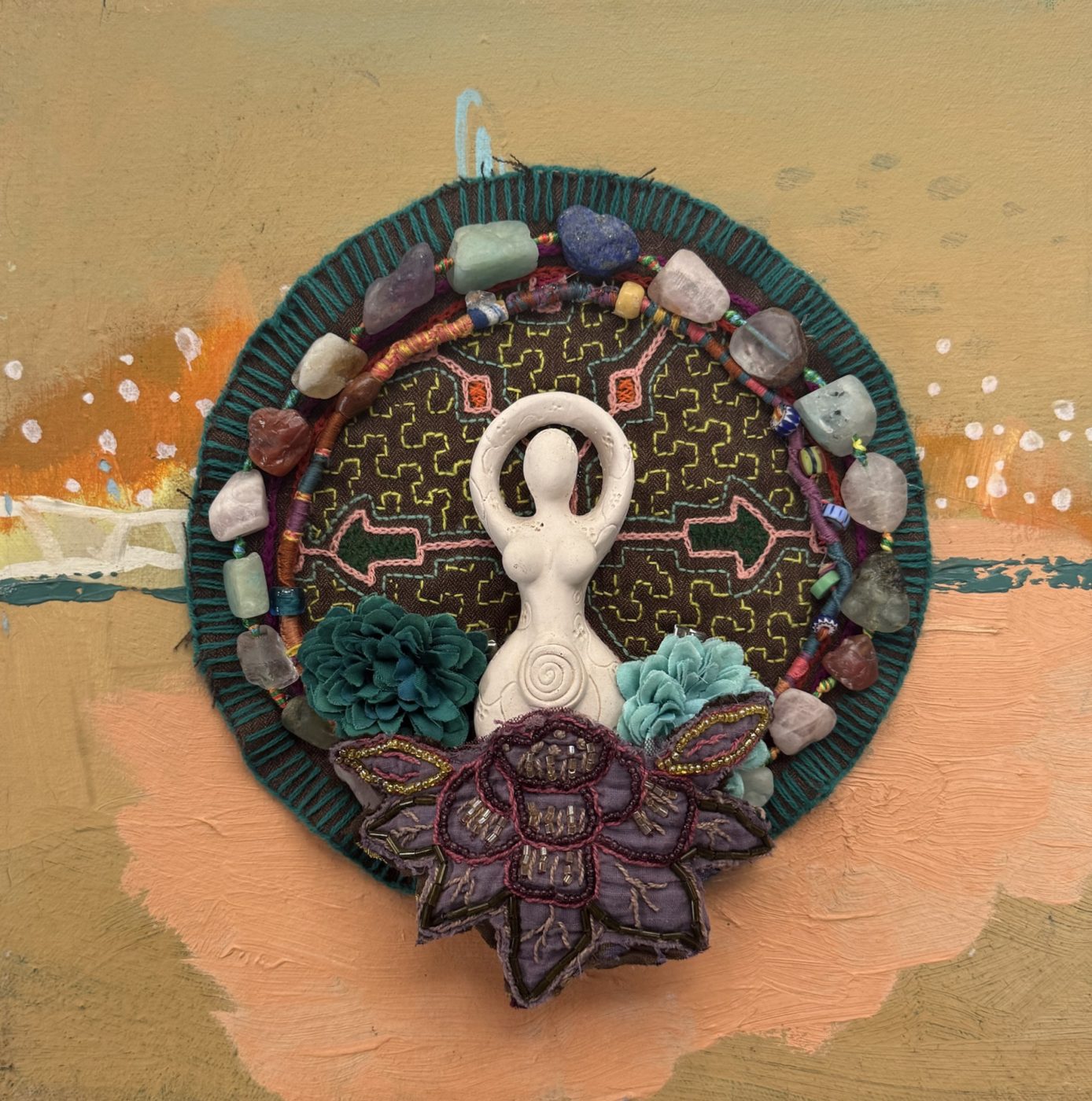 A white goddess figure on Shipibo-style embroidery, ringed with tumbled stones and wrapped cord, with fabric flowers and a beaded lotus, on a painted ochre and peach board.