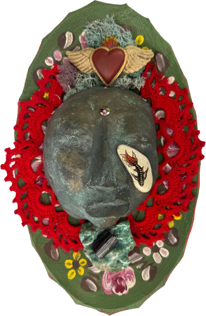 A dark sculpted face framed in red crochet on a green painted oval, with a winged sacred heart, lichen and a painted fish stone.