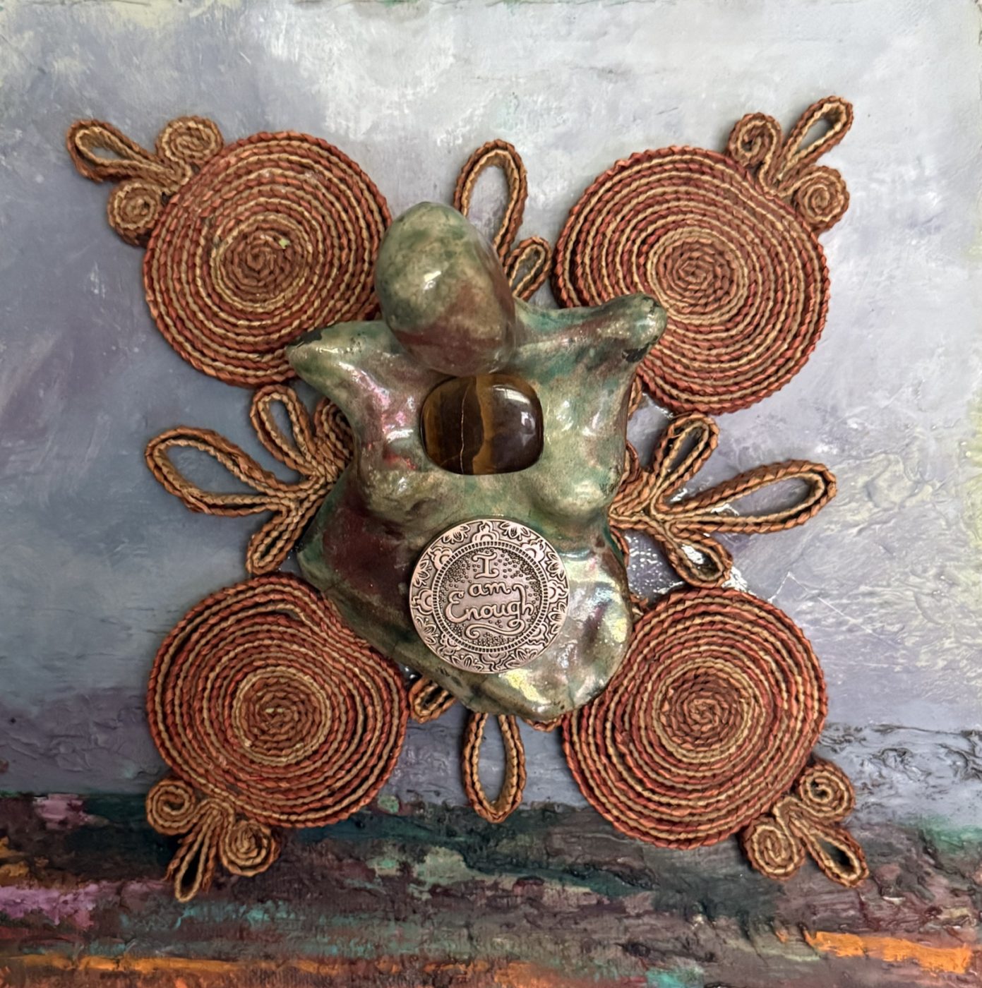 A raku goddess with a tiger’s eye stone and an I am Enough medallion, on coiled cord spirals over a painted lavender landscape.
