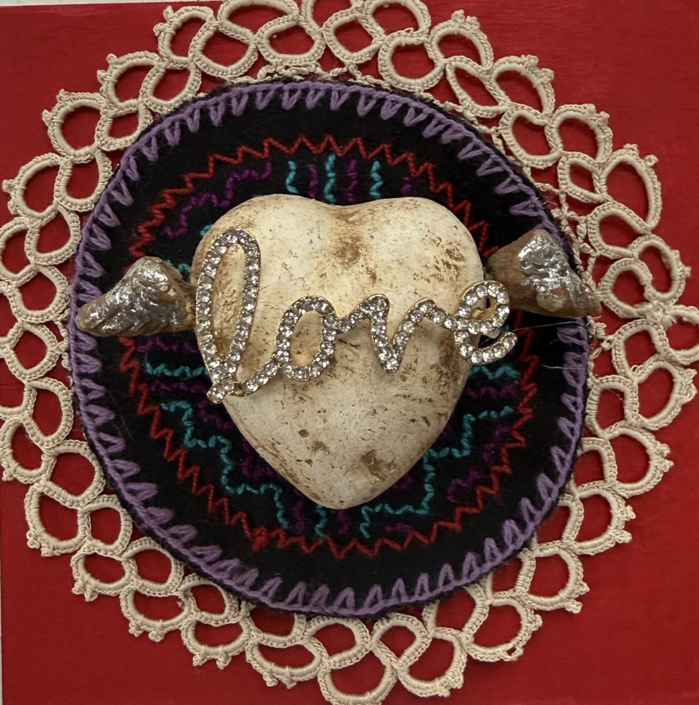 A winged stone heart with a rhinestone love script, on an embroidered black circle ringed in cream tatted lace, on a red board.