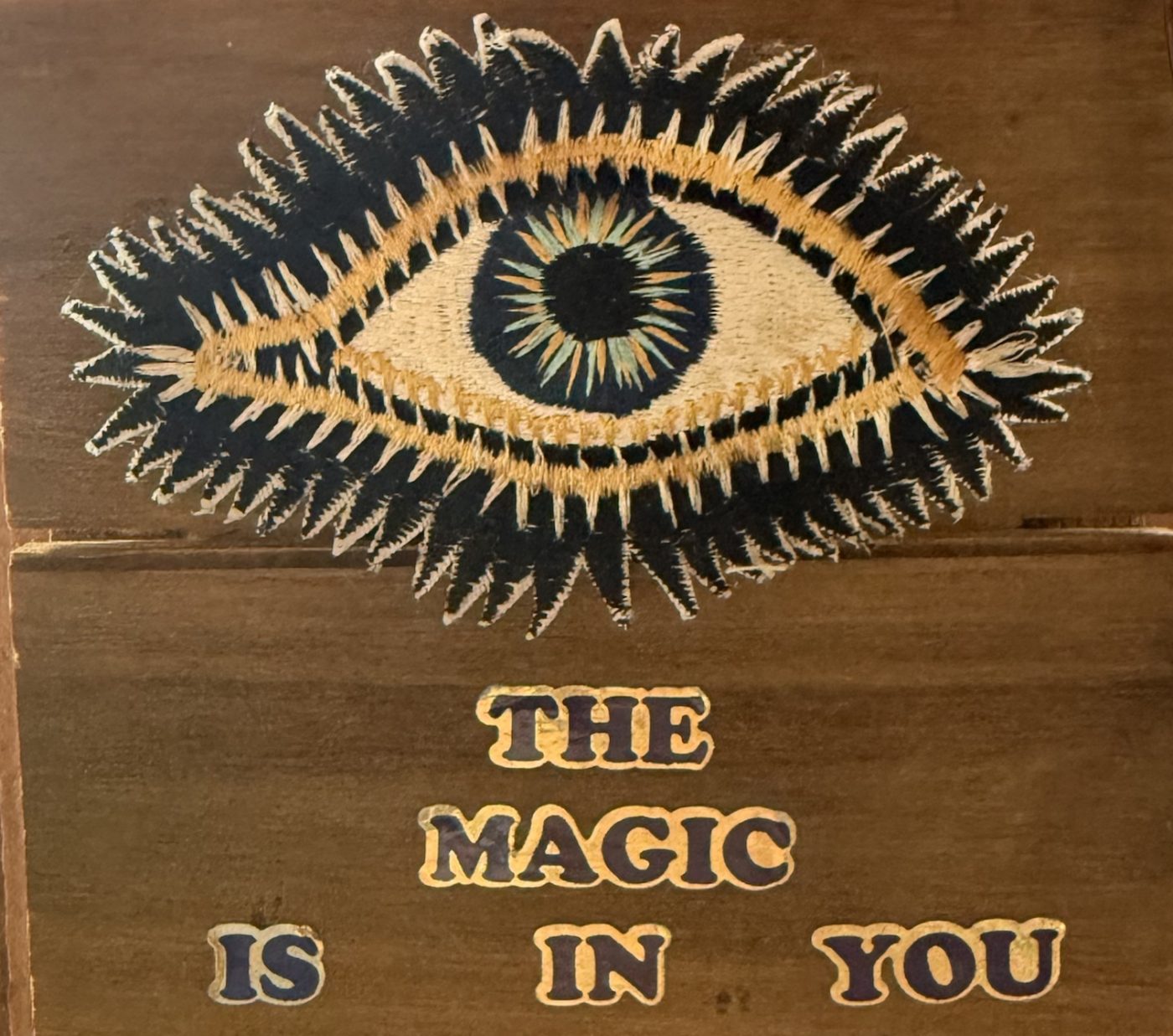 An embroidered eye with dark spiky lashes on weathered wood planks, above the words The Magic Is In You.