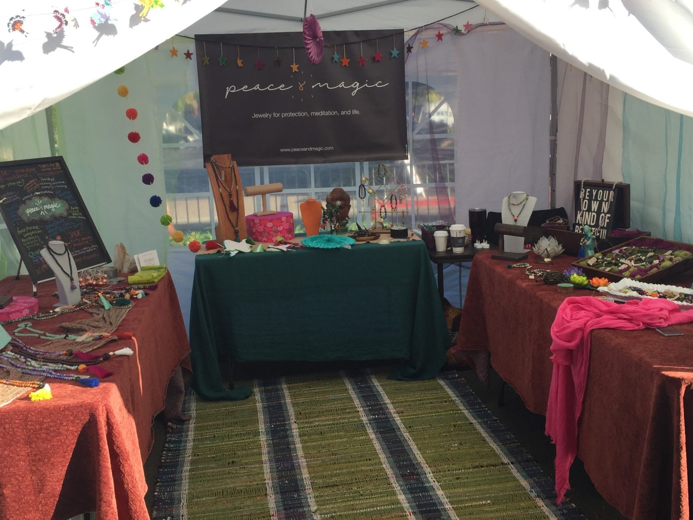 An outdoor Peace & Magic booth with tables of jewelry and a striped rug.