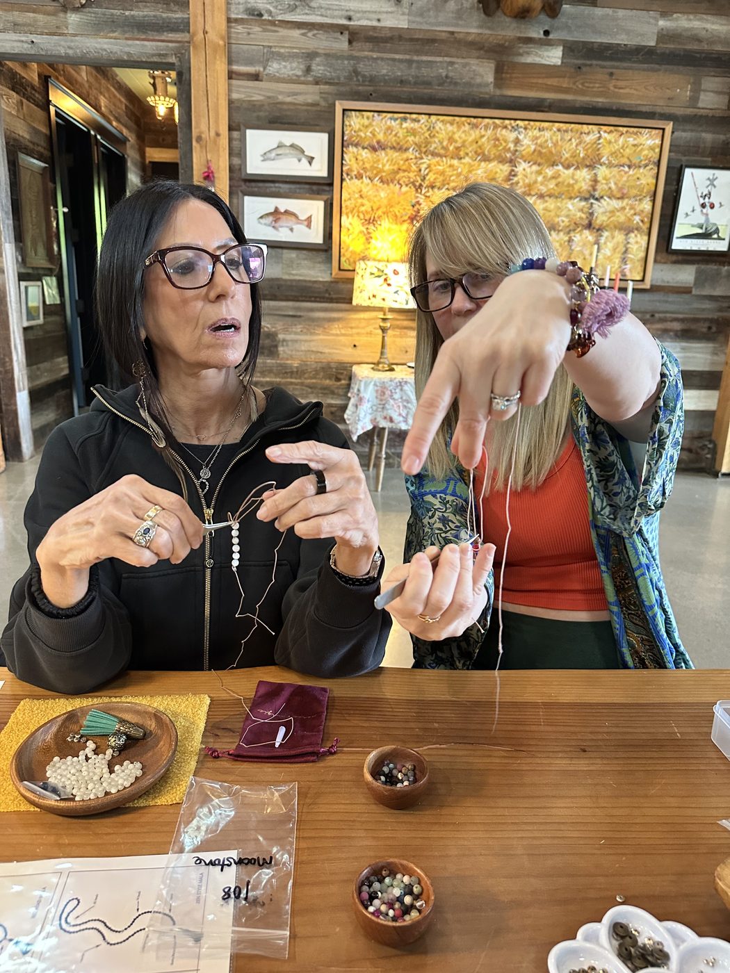 Amy teaching a woman to knot a mala at a wooden table, with bowls of beads.